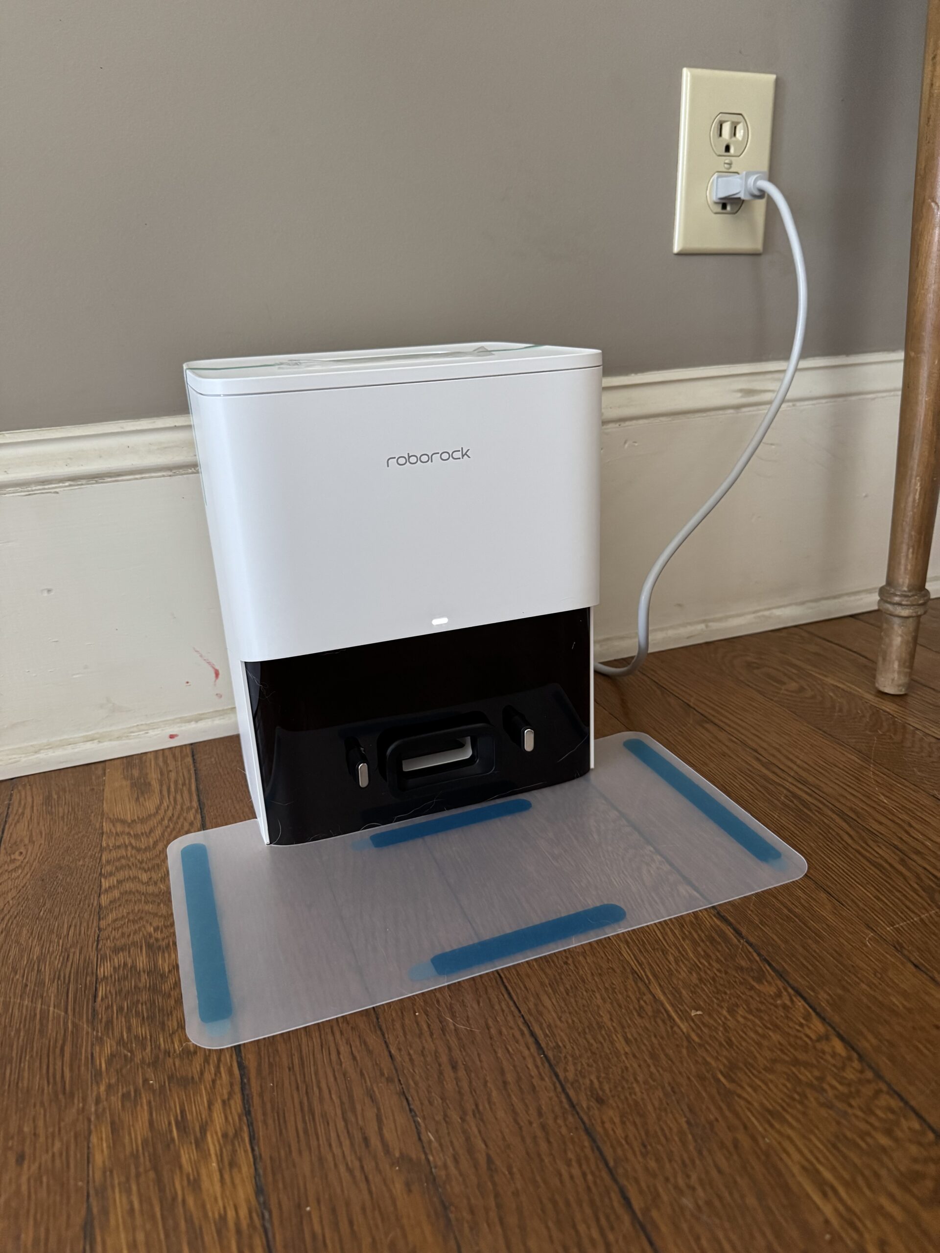 A Roborock charging station is plugged into a wall outlet and placed on a hardwood floor, sitting on a clear mat with blue strips.
