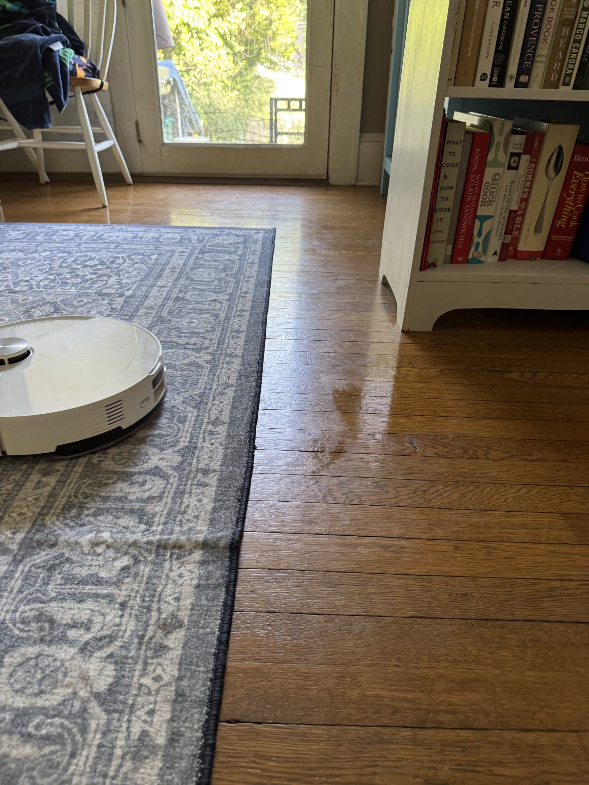 A white robot vacuum is cleaning the hardwood floor next to a patterned rug and a white bookshelf filled with books near a glass door.