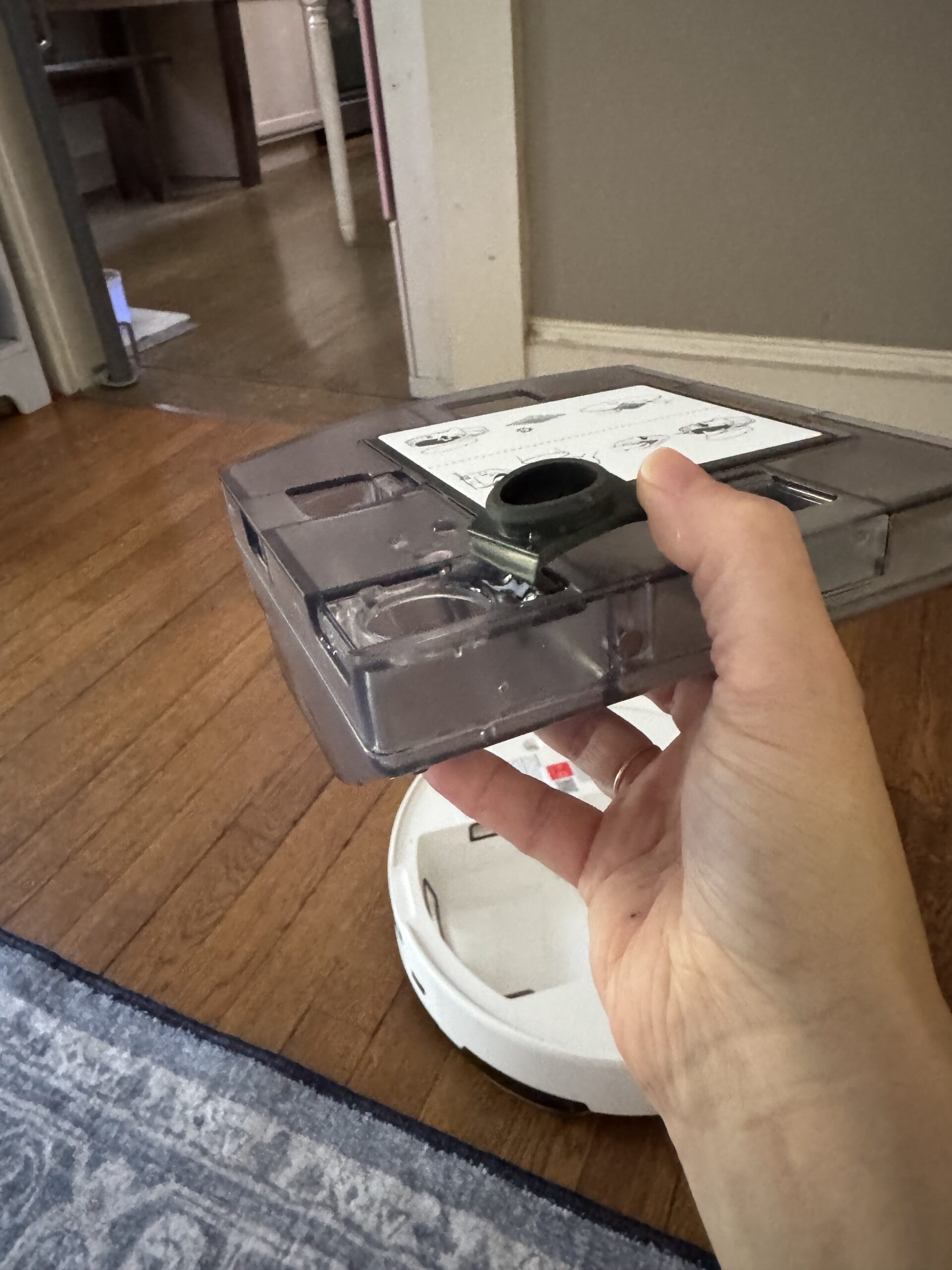 A hand holds a clear plastic dustbin from a robot vacuum, with the vacuum base on a wooden floor and a rug partially visible.