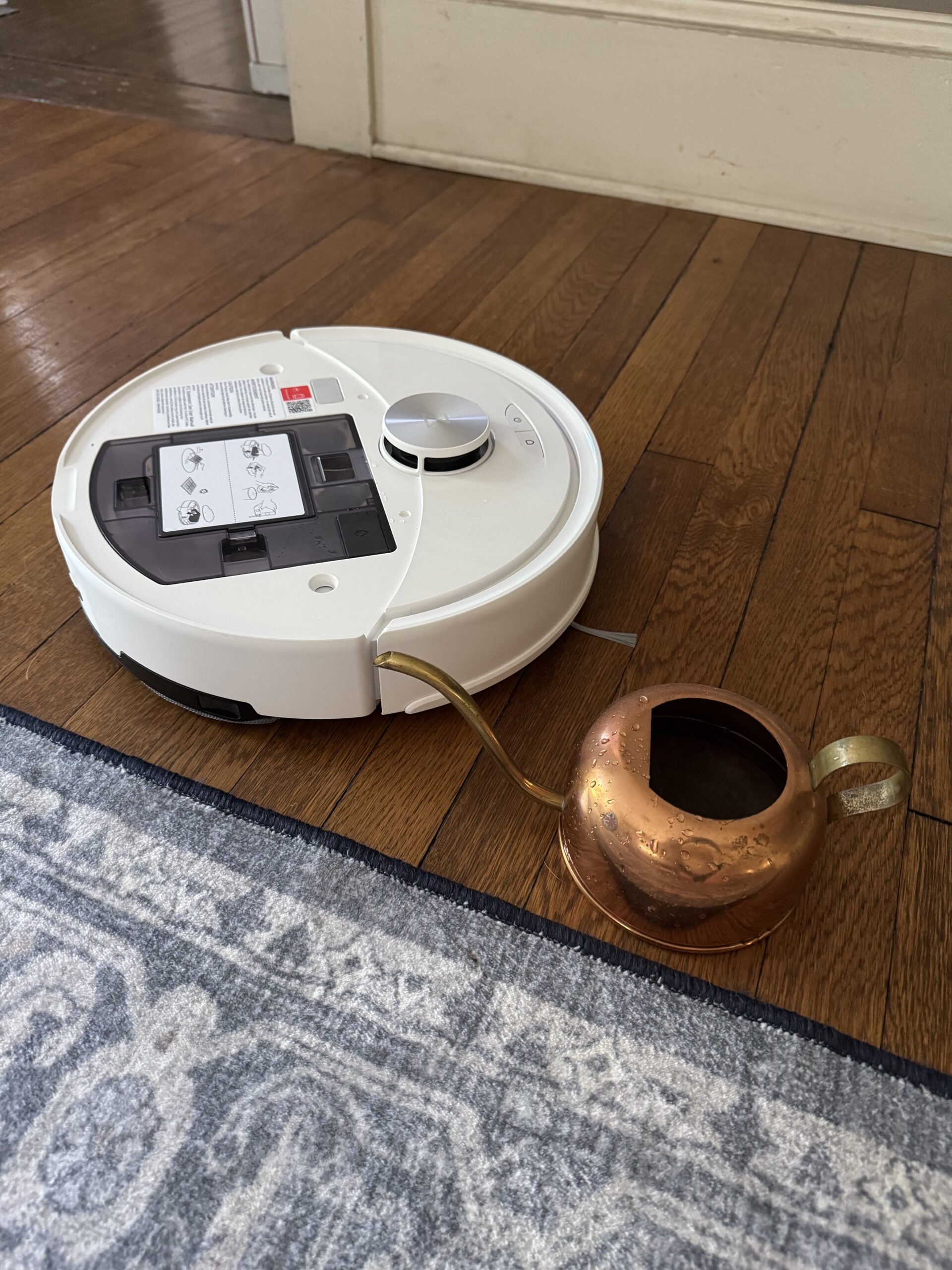 A white robotic vacuum cleaner on a wooden floor sits next to a copper watering can with a curved spout, near the edge of a blue and white patterned rug.