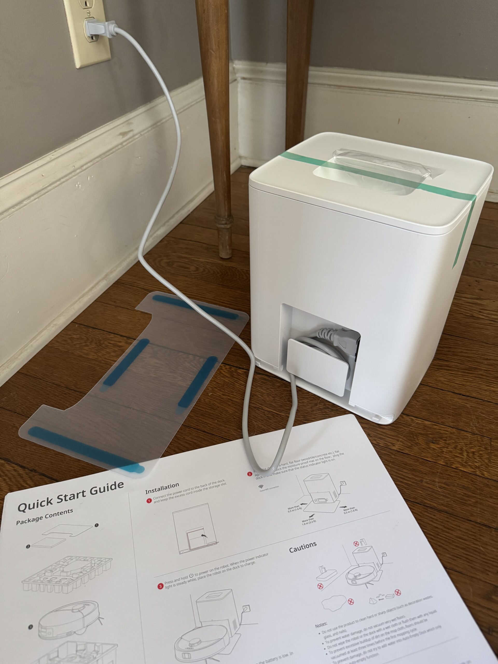 A white appliance plugged into a wall sits on a wooden floor beside a plastic mat and an open quick start guide with installation instructions.