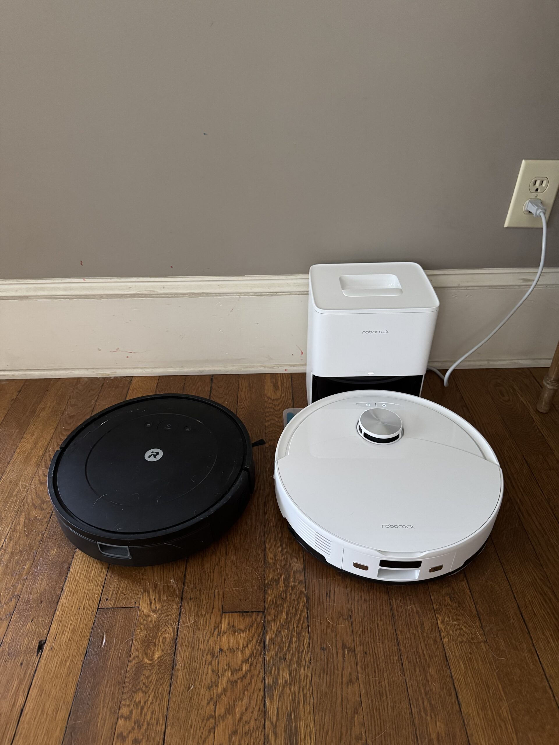 Two robot vacuum cleaners, one black and one white, are positioned on a hardwood floor near a charging station plugged into the wall.