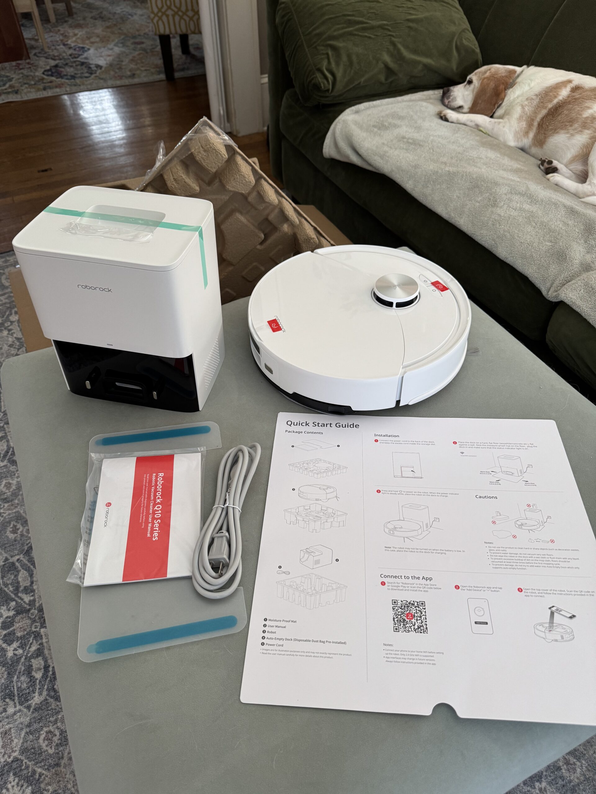 A robot vacuum and its charging dock are on a table with accessories, power cord, two blue strips, and a quick start guide. A dog is lying on a couch in the background.