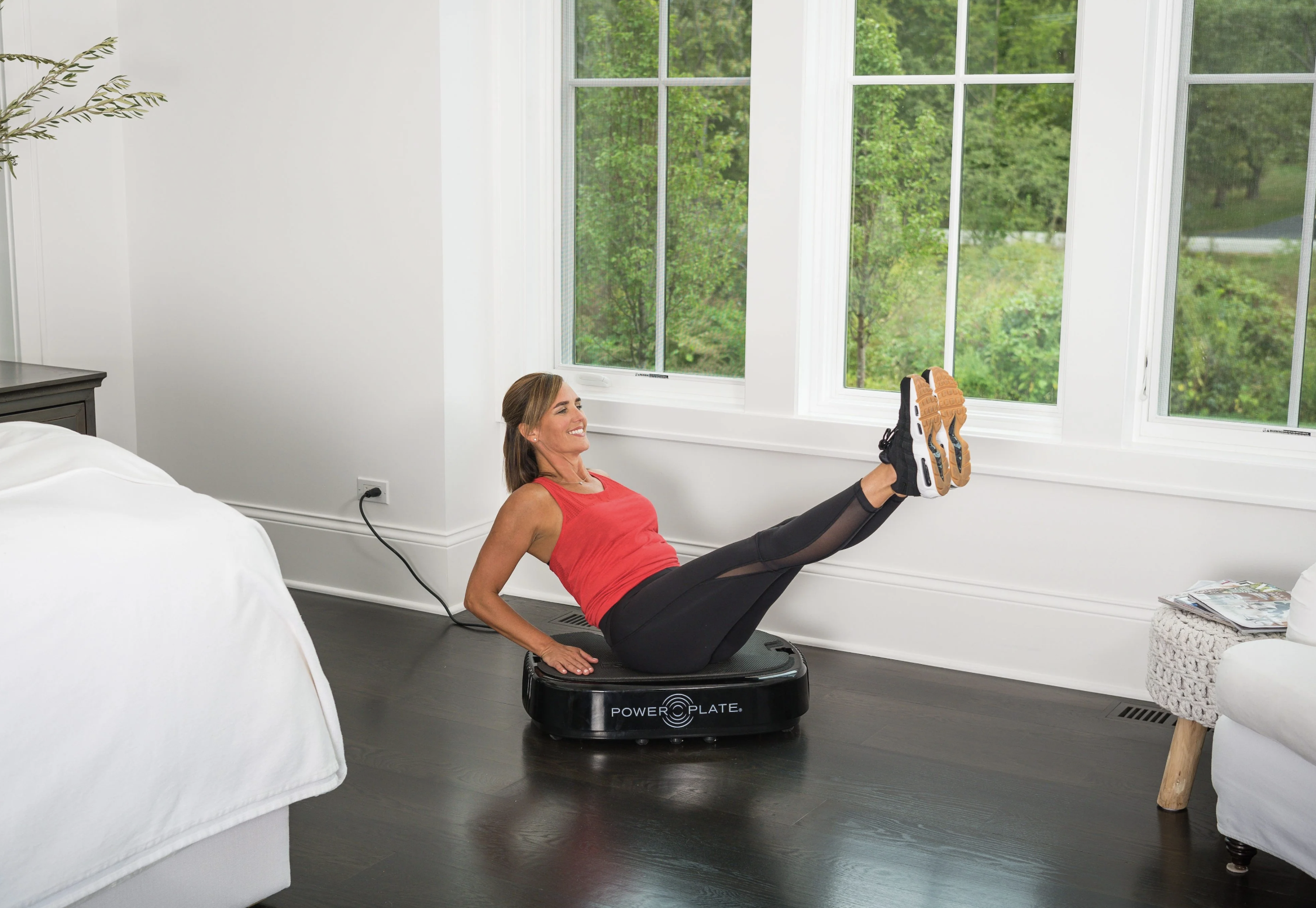 A woman in athletic clothing performs a leg raise exercise on a Power Plate vibration platform, with massage guns nearby, in a bright bedroom with large windows.