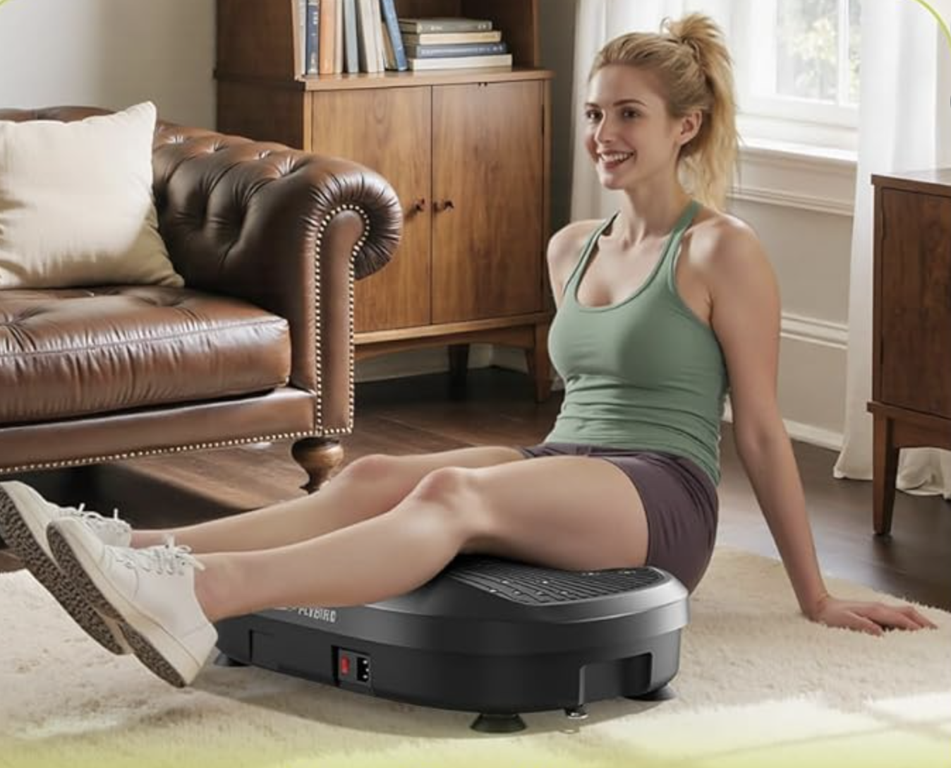 A woman in workout clothes sits on the floor with her legs resting on a black vibration plate, while massage guns sit nearby in the living room.