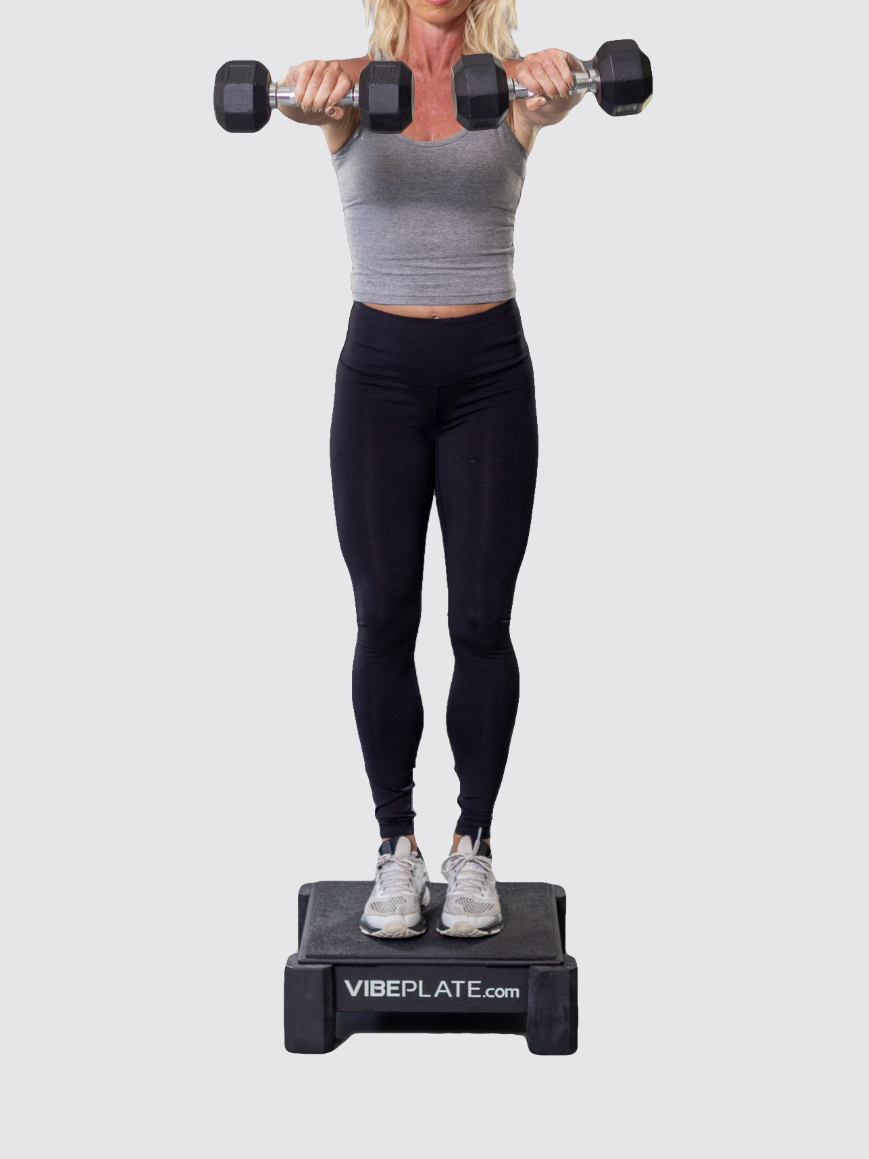 A person stands on a platform, holding dumbbells in both hands, arms extended forward at shoulder height, wearing athletic clothing—massage guns nearby for post-workout recovery.