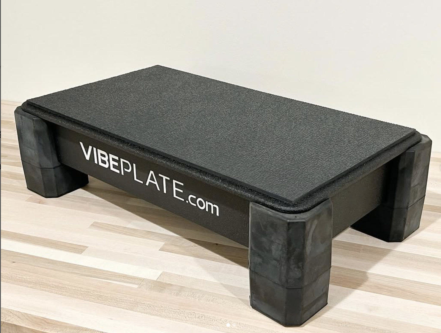 A black rectangular VibePlate exercise platform with four sturdy legs sits on a light wood floor against a plain wall, perfect for pairing with massage guns to enhance your recovery routine.