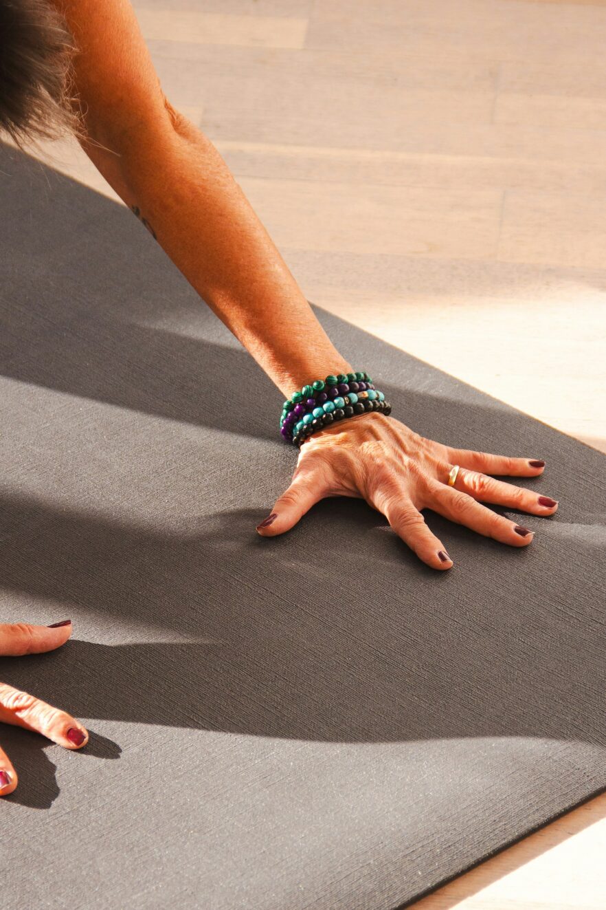 A person’s hand, wearing beaded bracelets, is pressed against a yoga mat on a wooden floor, suggesting a yoga or exercise pose—perfect for easing tension before using massage guns.
