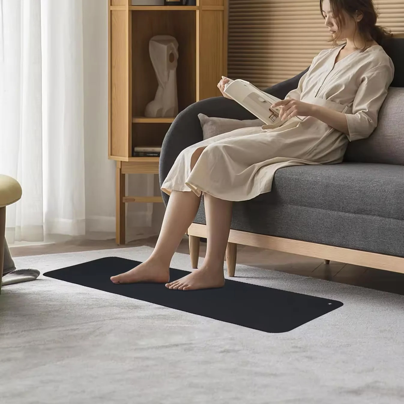 A woman in a light dress sits on a grey sofa reading a book, her bare feet resting on a black rectangular mat beside massage guns placed neatly on the light carpet.