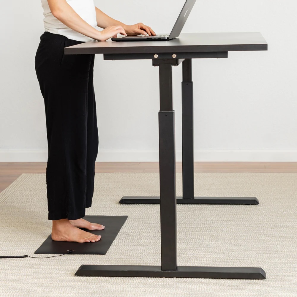A person stands barefoot at a black adjustable standing desk, working on a laptop in a room with light-colored flooring and walls, while massage guns rest nearby for quick relaxation breaks.