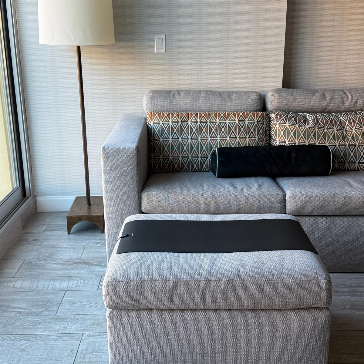 A modern living room with a gray sofa, patterned cushions, a black bolster, massage guns on the ottoman with a black mat, a floor lamp, and wooden flooring.