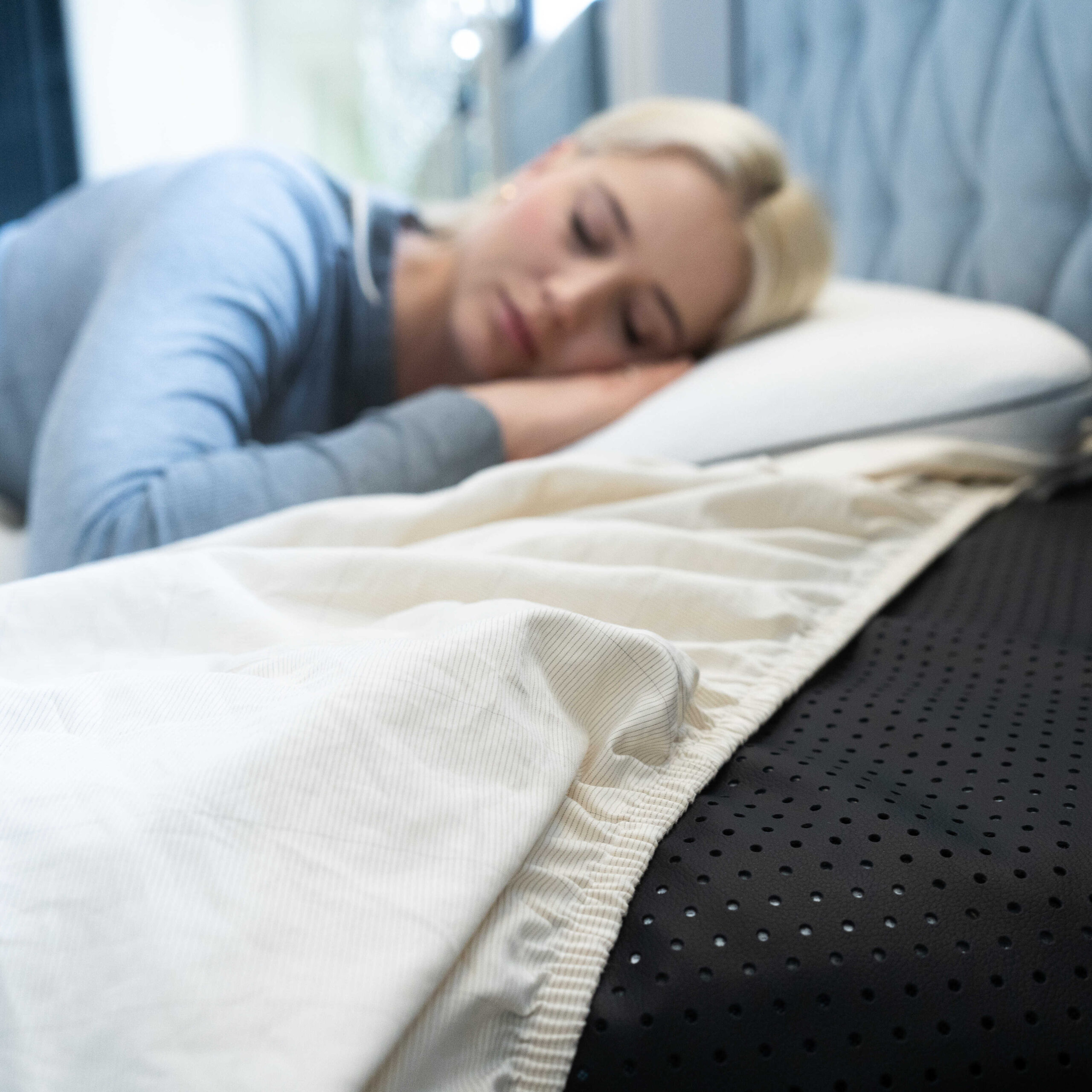 A woman with blonde hair sleeps on her side in bed, resting her head on a white pillow with a cream-colored blanket and a black perforated mattress cover visible—massage guns rest nearby for ultimate relaxation.
