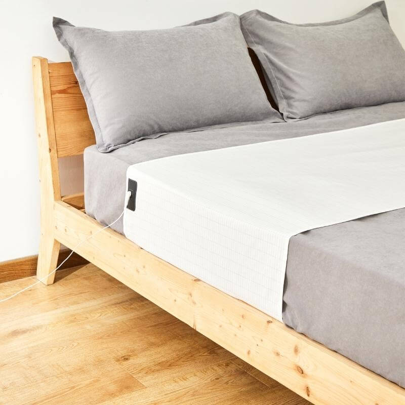 A wooden bed frame with gray bedding and pillows, featuring a white electronic heating pad placed across the lower half of the mattress—perfect for relaxing after using massage guns.