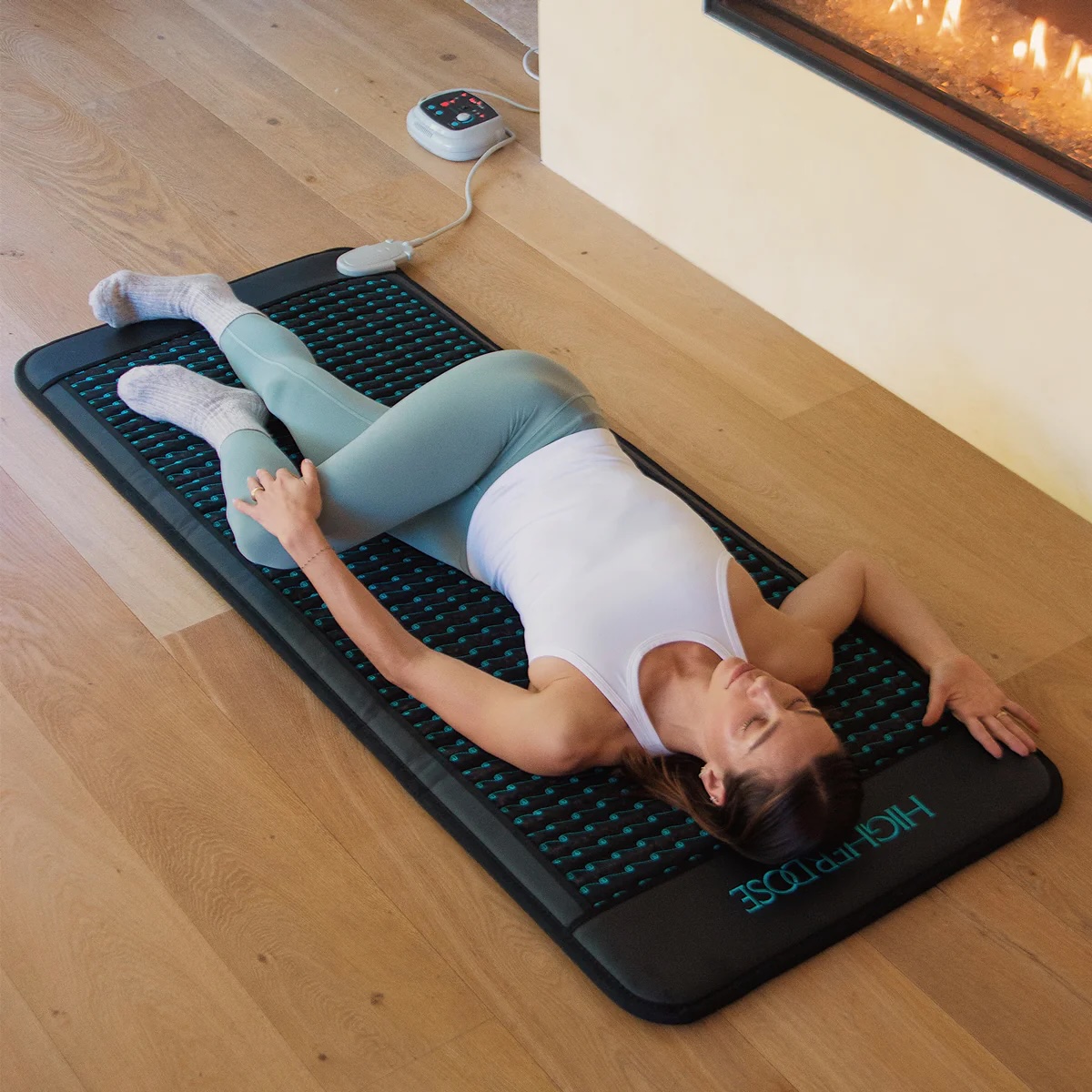 Woman lying on a heated mat with a remote control, stretching her leg on a wooden floor near a fireplace, with massage guns nearby for added relaxation.