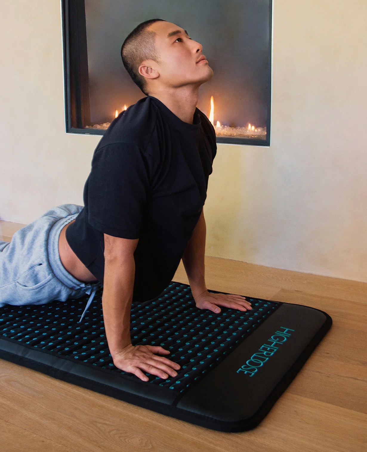A person performs a yoga stretch on a black acupressure mat in front of a modern fireplace, wearing a black shirt and gray sweatpants, while massage guns rest nearby for post-workout recovery.