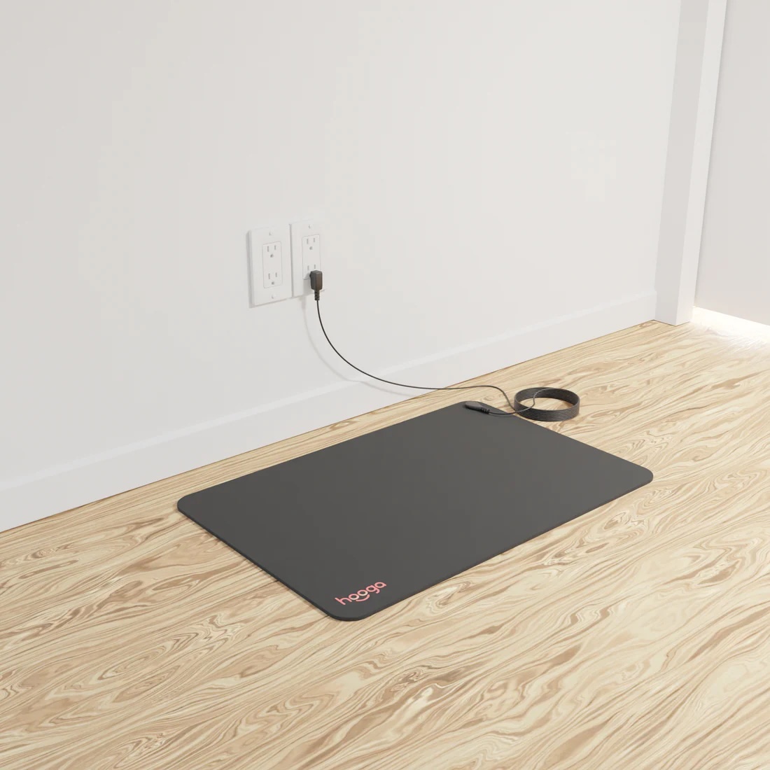 A rectangular black floor mat, perfect for use with massage guns, is plugged into a wall outlet and sits on a light wood floor near a white wall.
