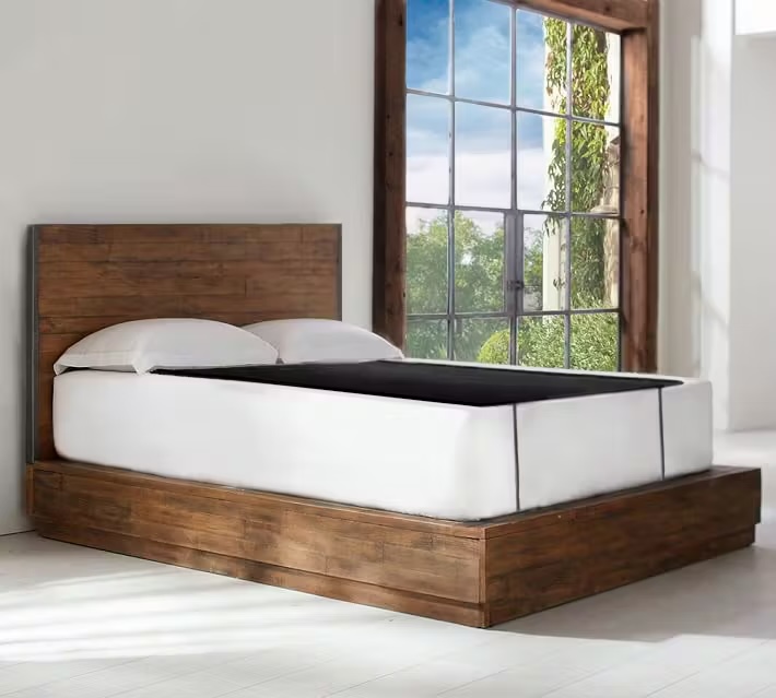 A wooden platform bed with a white mattress and pillows is positioned near a large window with a view of greenery outside, creating the perfect spot to unwind after using massage guns.