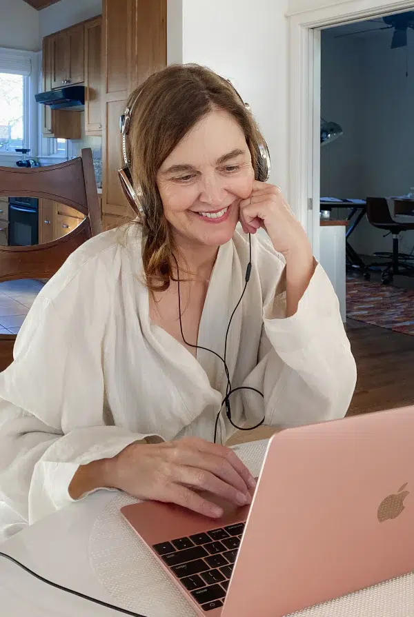 A woman wearing headphones and a light-colored robe sits at a table, smiling while using a pink laptop in a home setting.