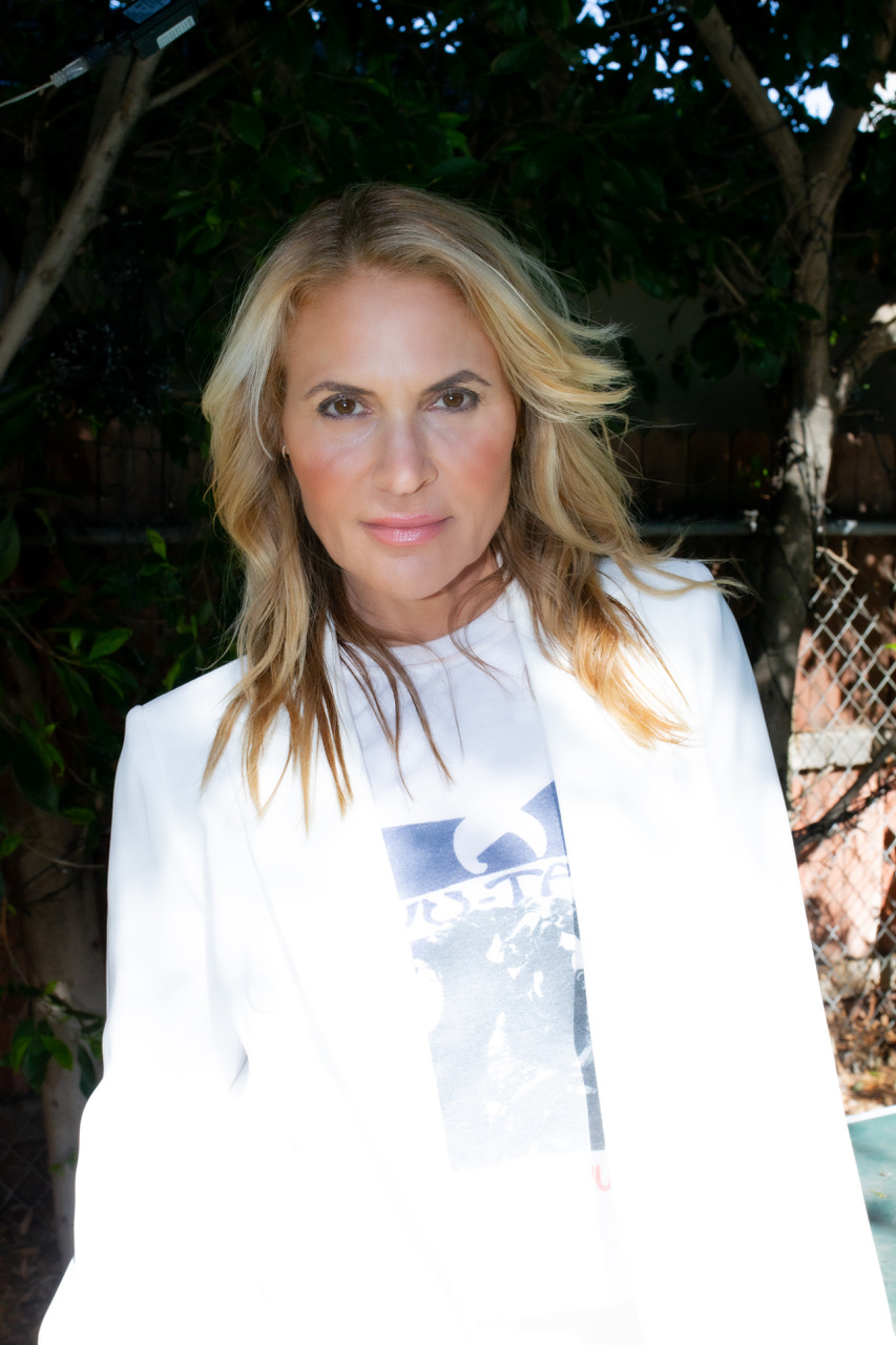 A woman with long blonde hair stands outdoors in partial sunlight, wearing a white blazer over a printed T-shirt. Trees and a fence are visible in the background.