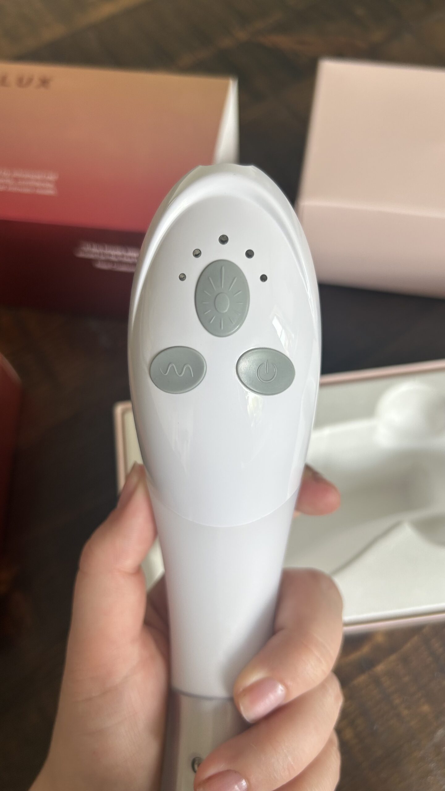 A hand holds a white handheld electronic device with three buttons and indicator lights, next to an open box with a molded compartment.