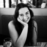 A woman with long dark hair sits at a table, resting her head on her hand and smiling at the camera. A glass of water is on the table in front of her.