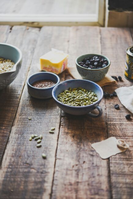 A wooden table displays bowls of pumpkin seeds, flaxseeds, dried fruit, a stick of butter, and a large bowl partially filled with oats.