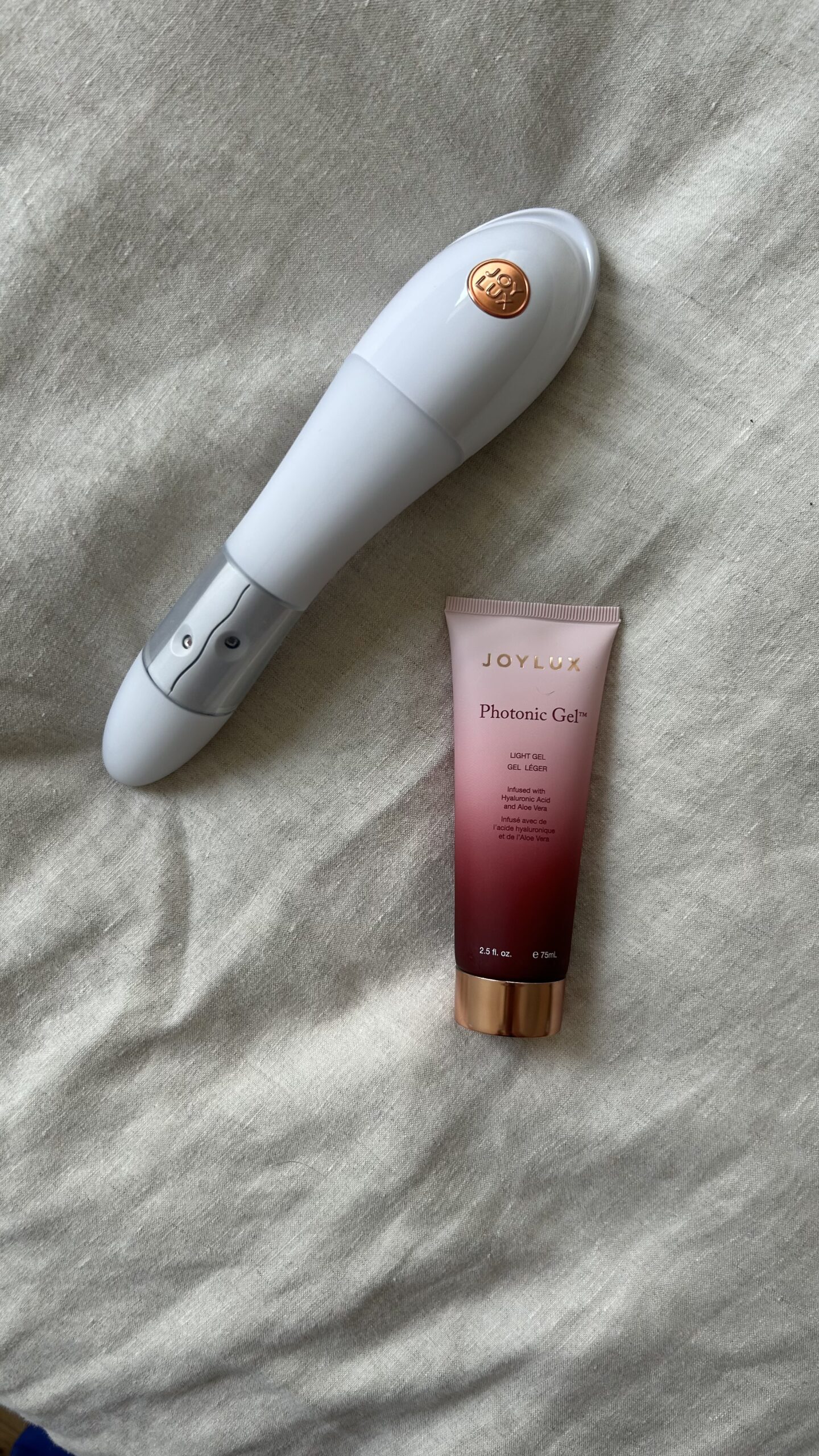 A white handheld electronic device and a tube of JOYLUX Photonic Gel are placed on a beige fabric surface.