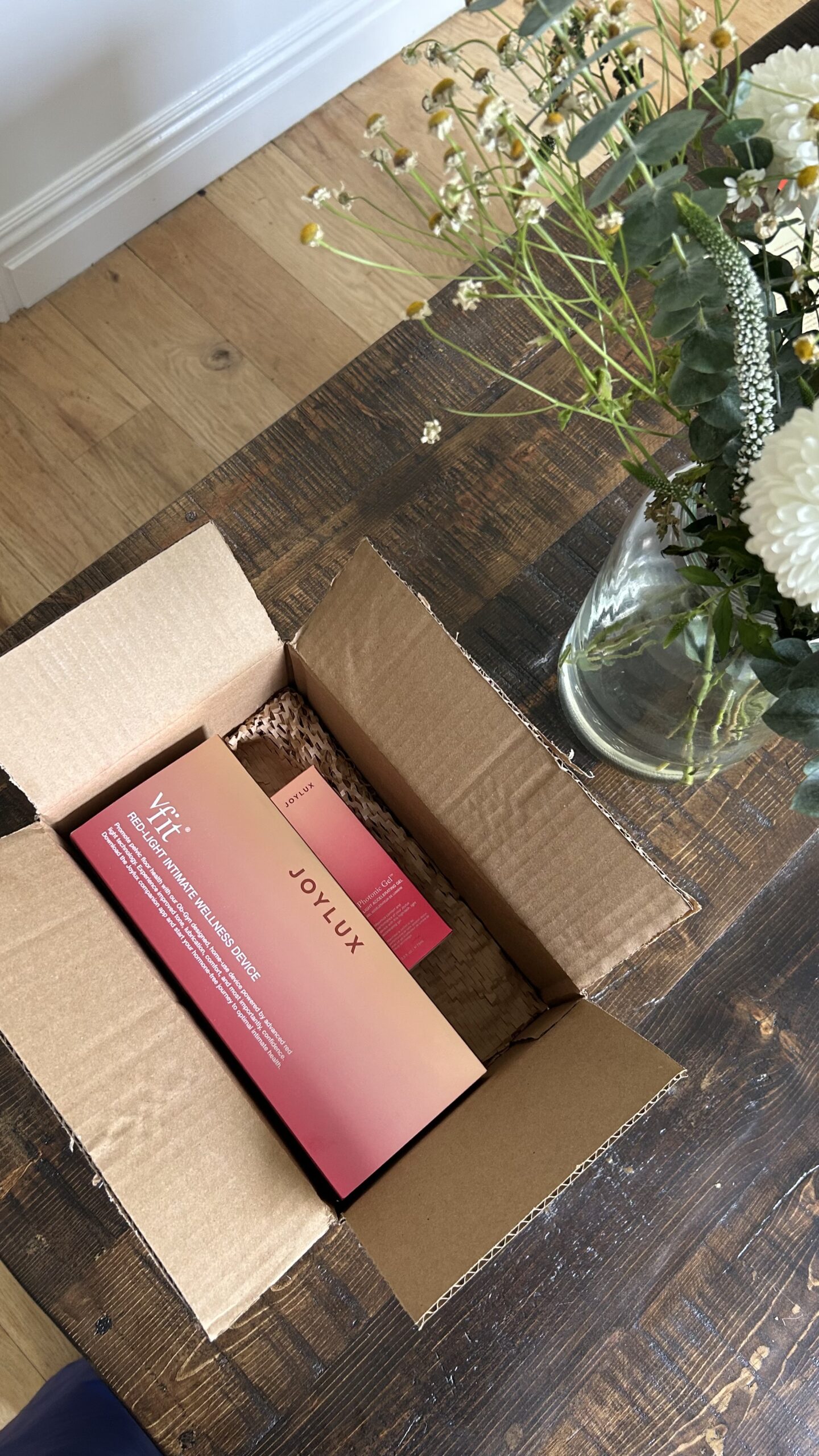 An open cardboard box on a wooden table contains a pink and orange Joylux product package next to a vase of white flowers.