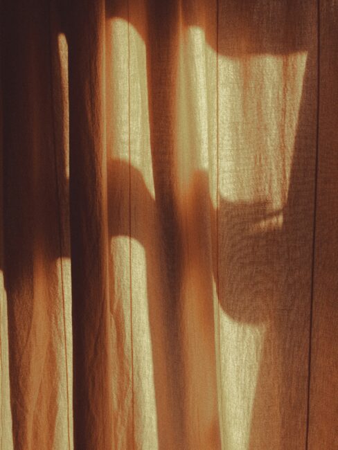 Sunlight casts soft, warm shadows through a window onto beige curtains, creating abstract patterns of light and shadow on the fabric.