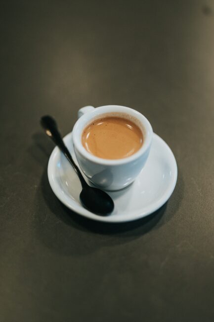 A white cup of espresso sits on a matching saucer with a black spoon, placed on a dark surface.