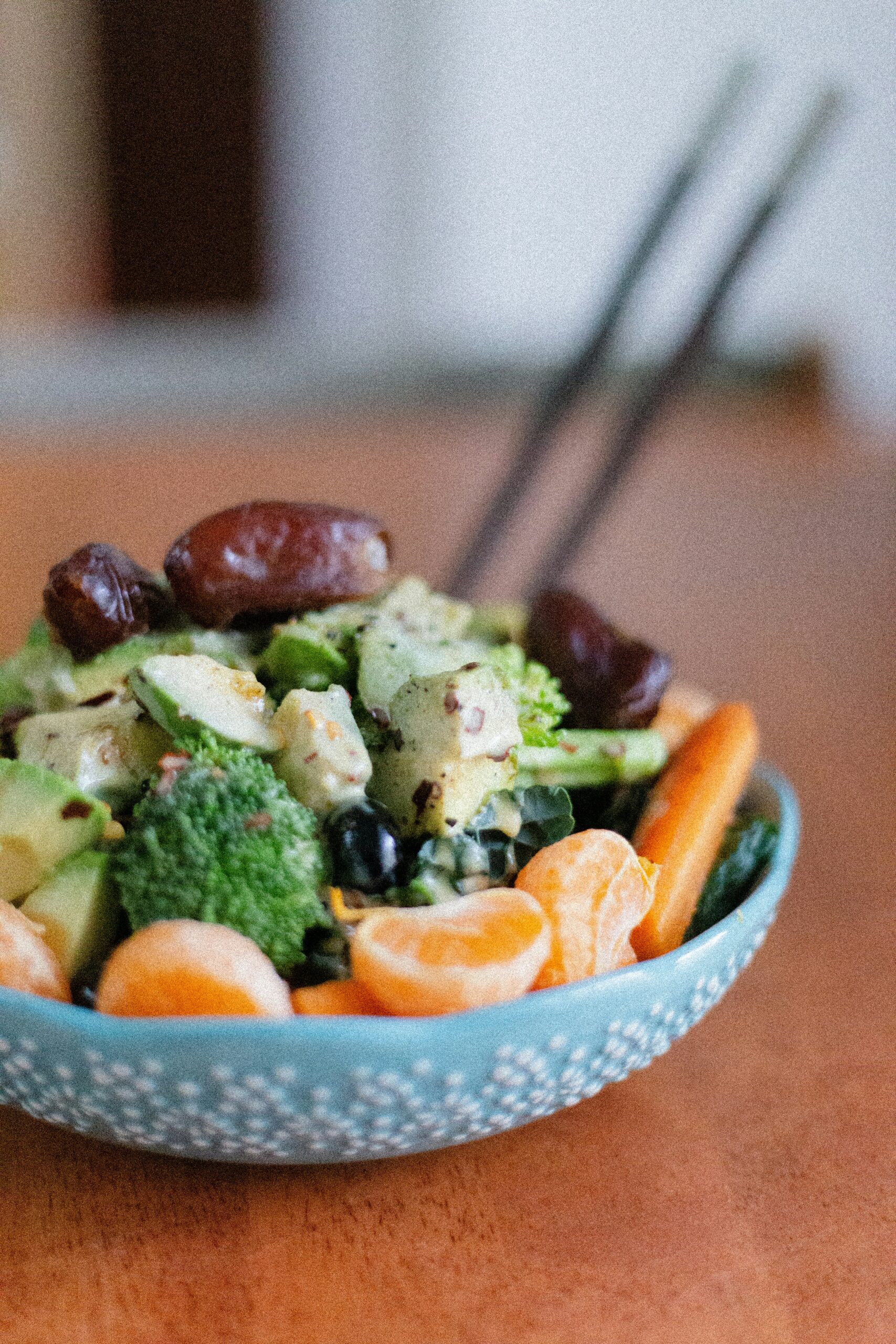 A bowl of salad with broccoli, avocado, olives, dates, carrots, and orange slices, with chopsticks resting on the bowl.
