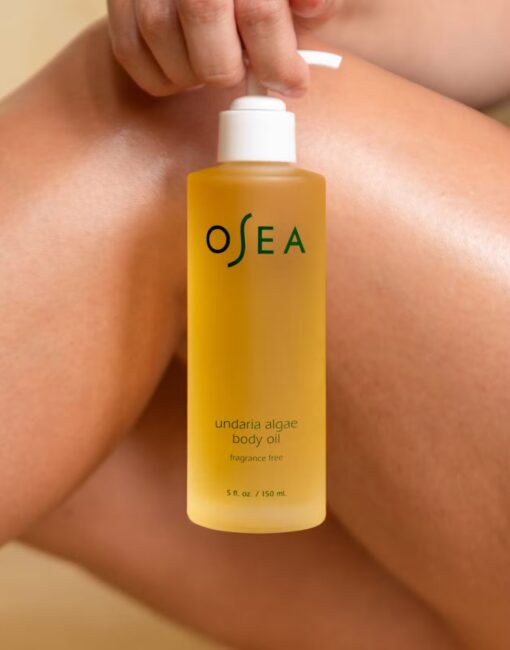 A person holds a pump bottle of OSEA Undaria Algae Body Oil against their legs. The bottle contains 150 mL of fragrance-free oil.
