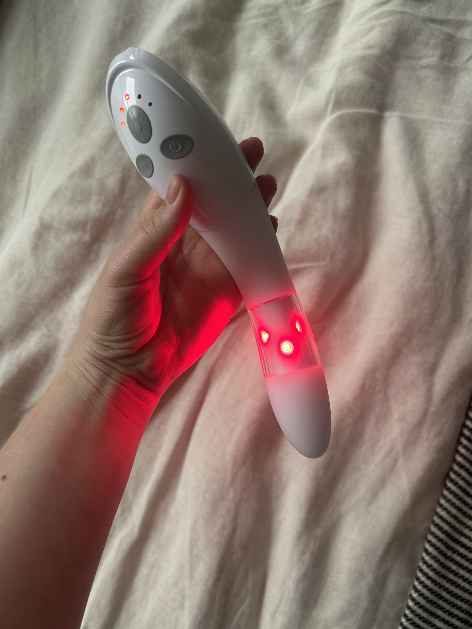 A hand holds a white handheld device emitting red LED lights, positioned over a cream-colored fabric background.