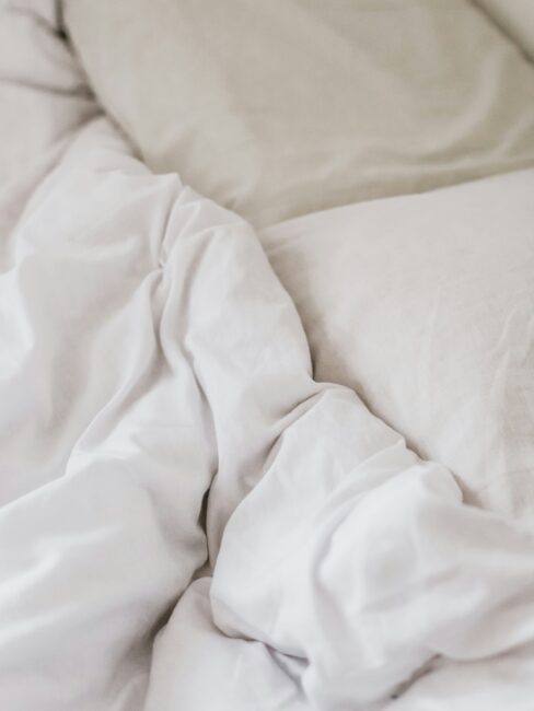 A close-up of unmade white bed sheets and a pillow, showing soft fabric folds and a relaxed, casual appearance.