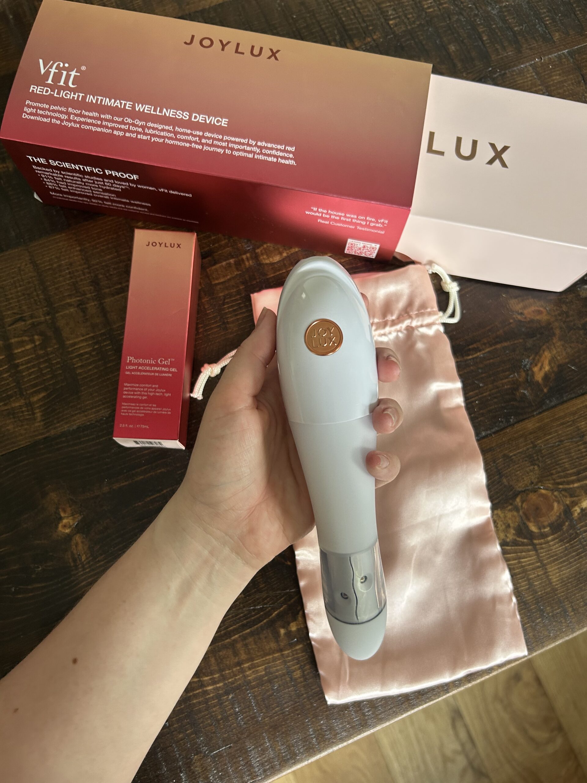 A hand holds a Joylux VFit red-light intimate wellness device, with its box, a small bottle, and a pink satin pouch on a wooden surface.