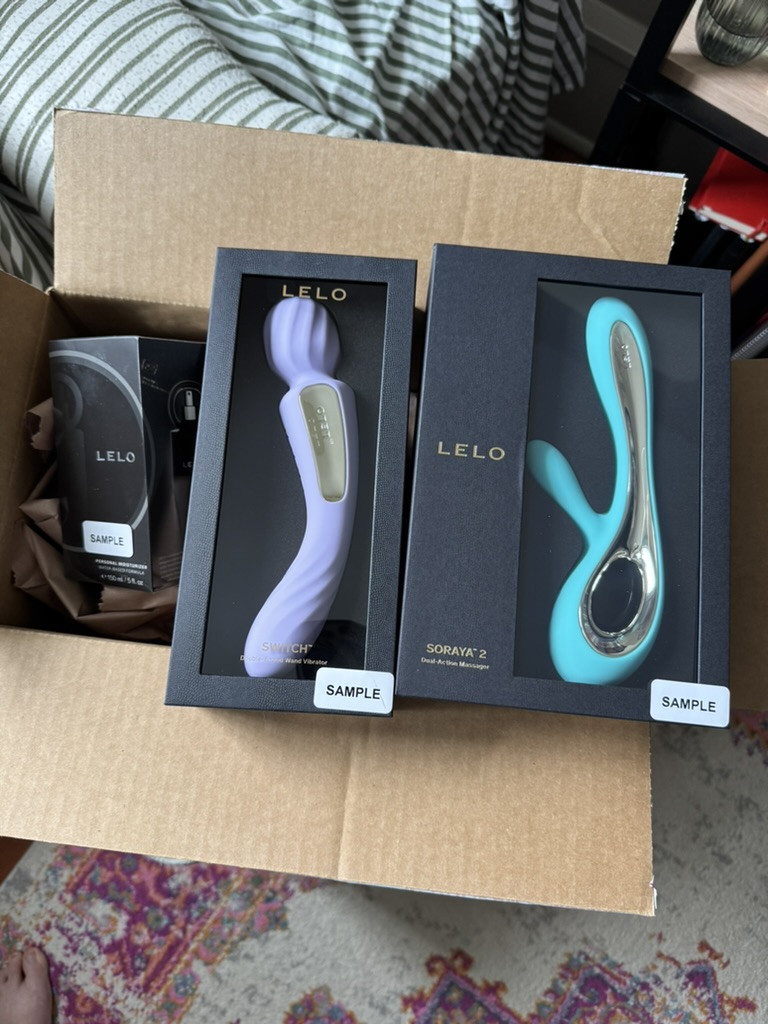 An open box contains two LELO branded personal massagers in black packaging, labeled as samples, with one device in lavender and another in teal color.