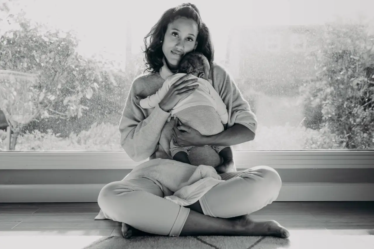 A woman sits cross-legged on the floor, holding and embracing a baby in her arms, in front of a large window.