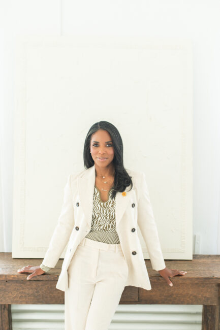A woman with long dark hair wearing a light-colored suit and patterned top stands in front of a large textured white canvas, leaning back on a wooden table.