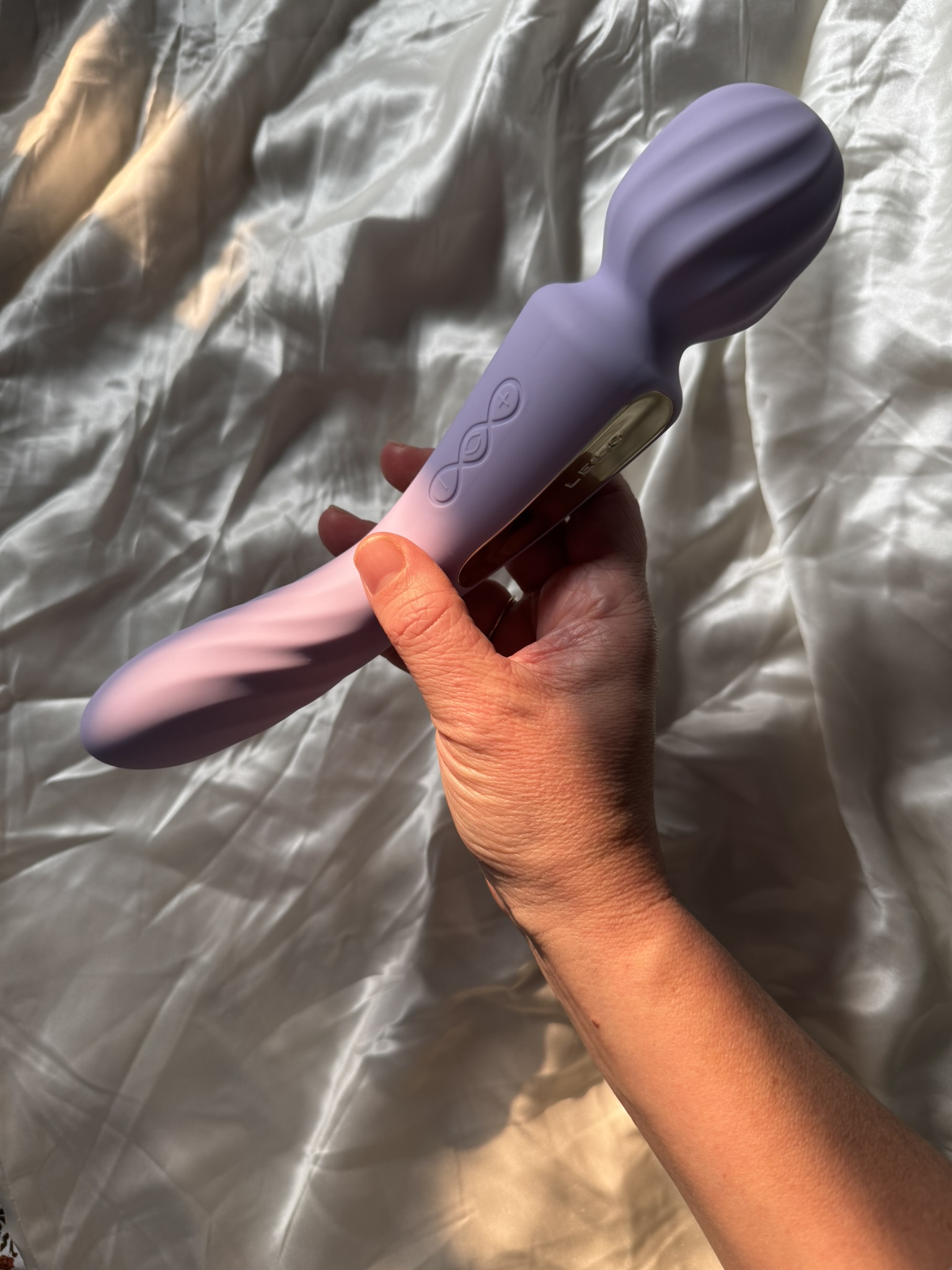 A hand holding a lavender-colored personal massager with control buttons, against a satin fabric background.