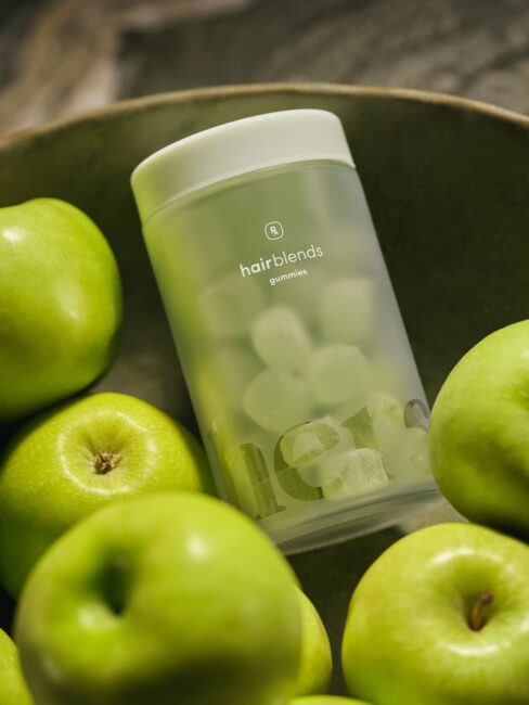 A frosted container labeled "hairblends gummies" surrounded by several green apples.