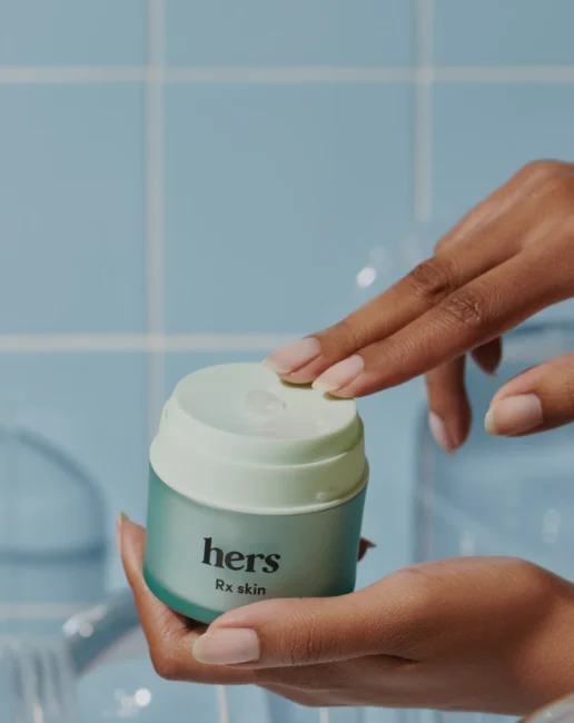 A person dispenses cream from a green and white "hers Rx skin" container with their finger, against a blue tiled background.