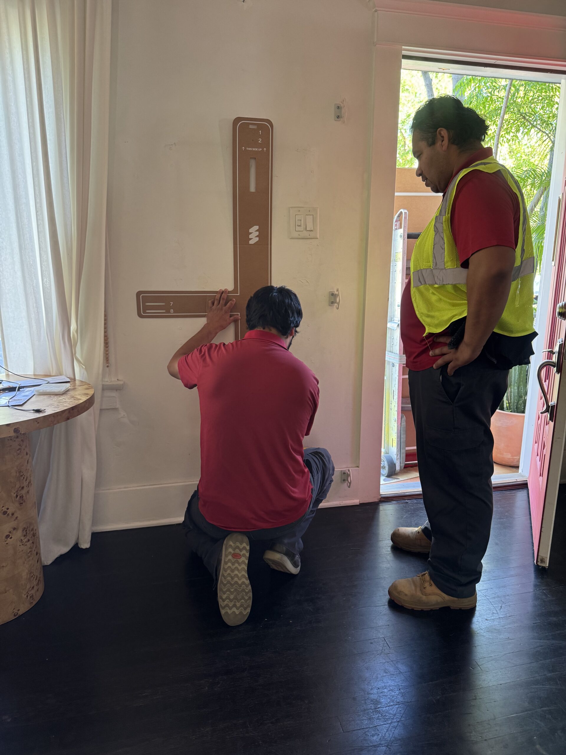 Two workers in red shirts, one kneeling and one standing, install a large L-shaped cardboard template on a white wall inside a room with dark floors and a window.