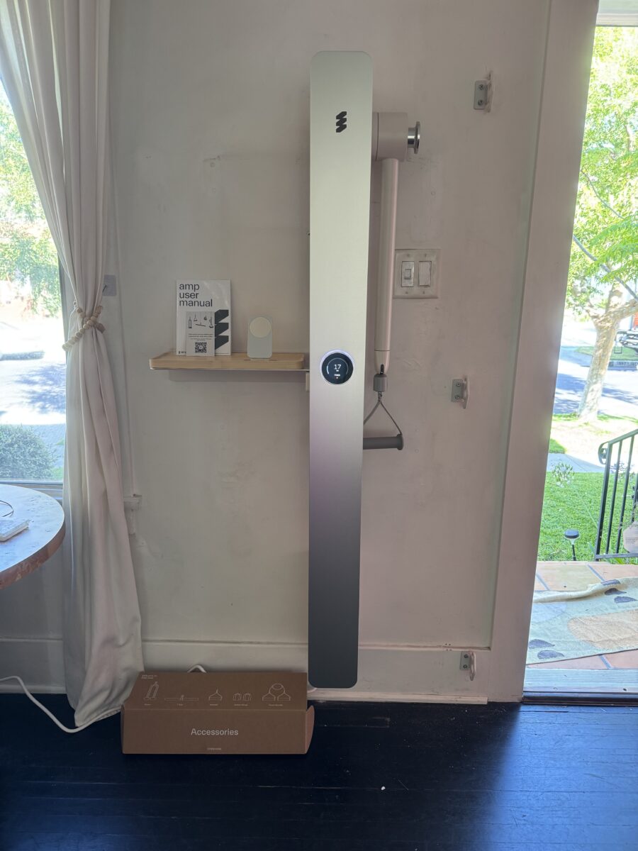 A tall, rectangular device is mounted on a wall beside a small shelf with a manual, and a cardboard box labeled "Accessories" sits on the floor below. A window is to the right.