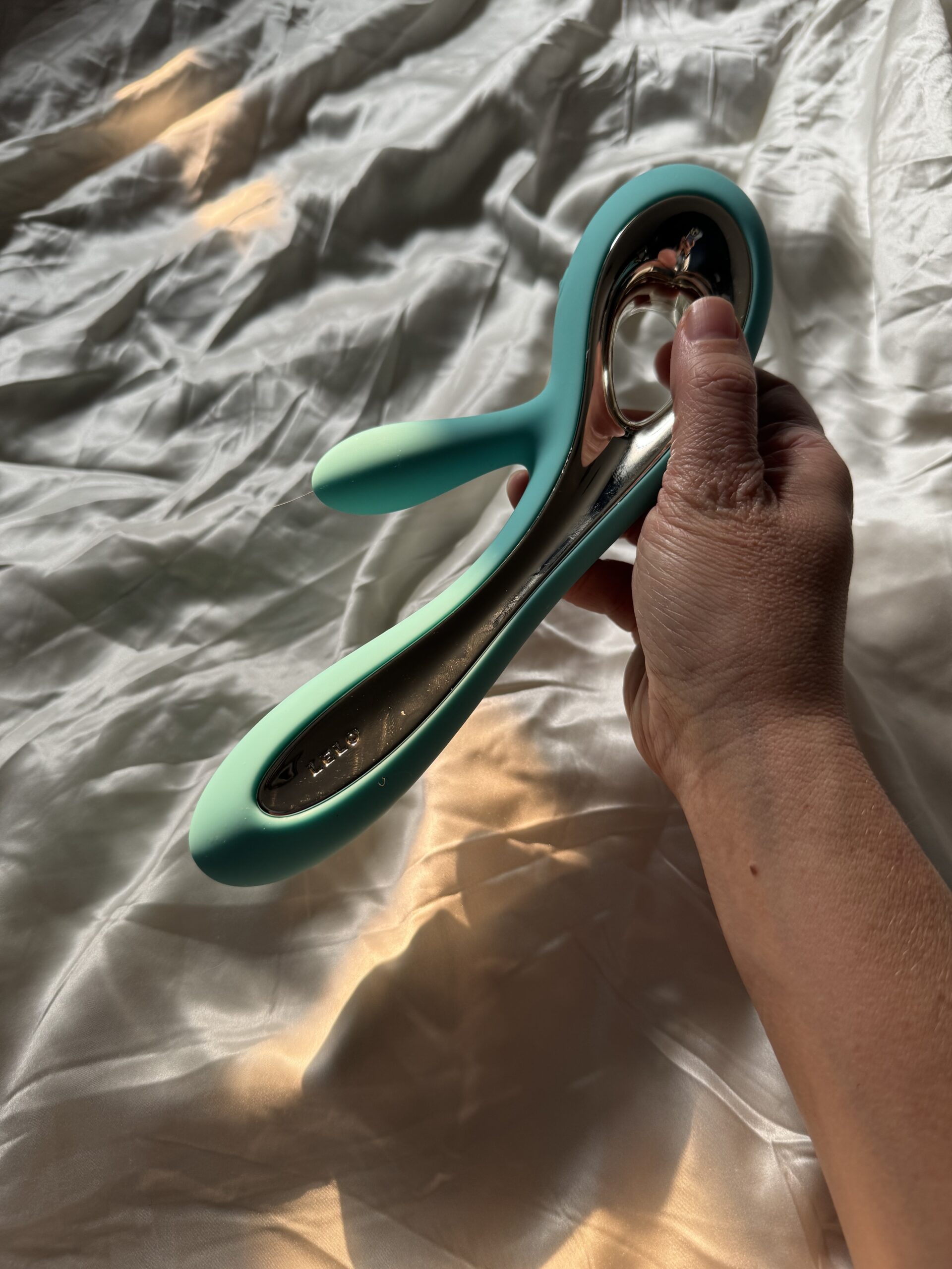 A hand holding a teal and silver silicone personal massager on a white, rumpled bedsheet.
