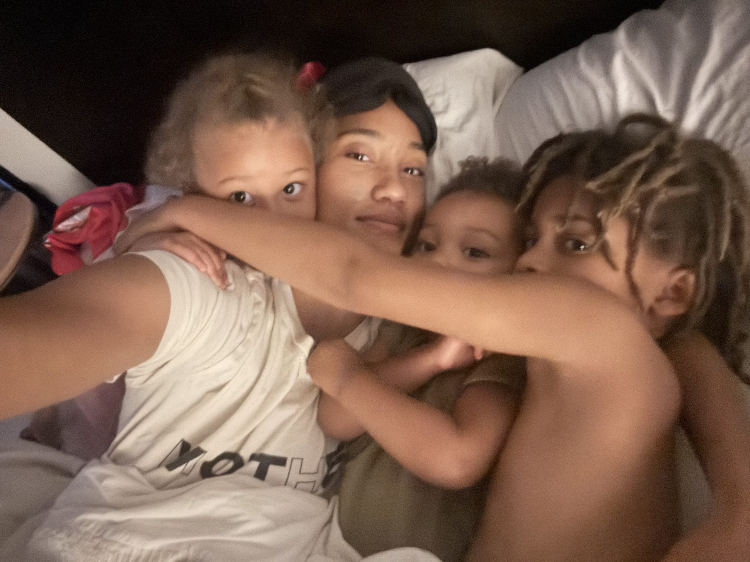 A woman and three children are lying close together on a bed, looking at the camera. One child is wearing a white shirt, and another has their arm around the others.