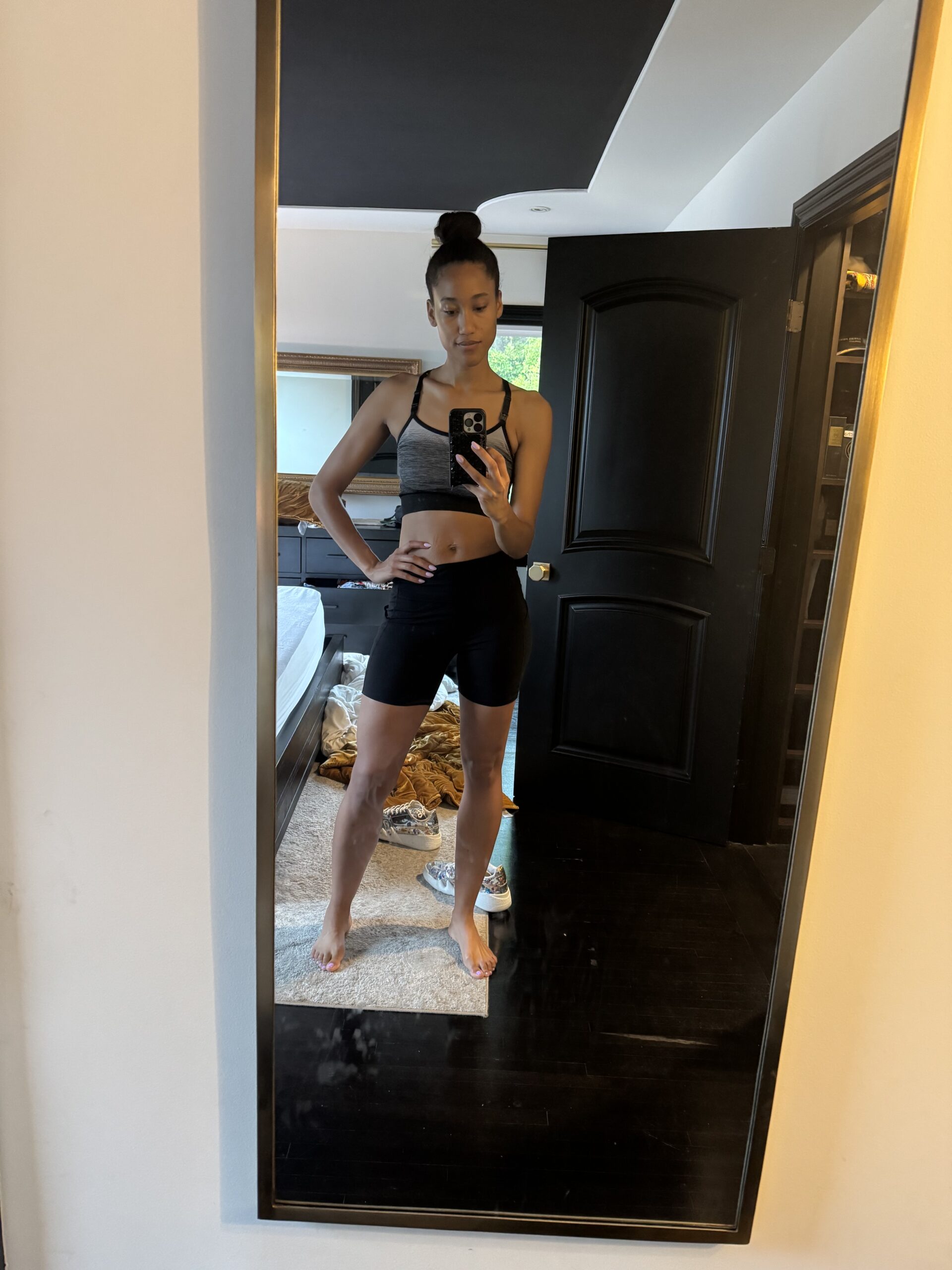 Woman taking a mirror selfie in a bedroom, wearing a sports bra and black shorts, standing barefoot on a carpeted floor.