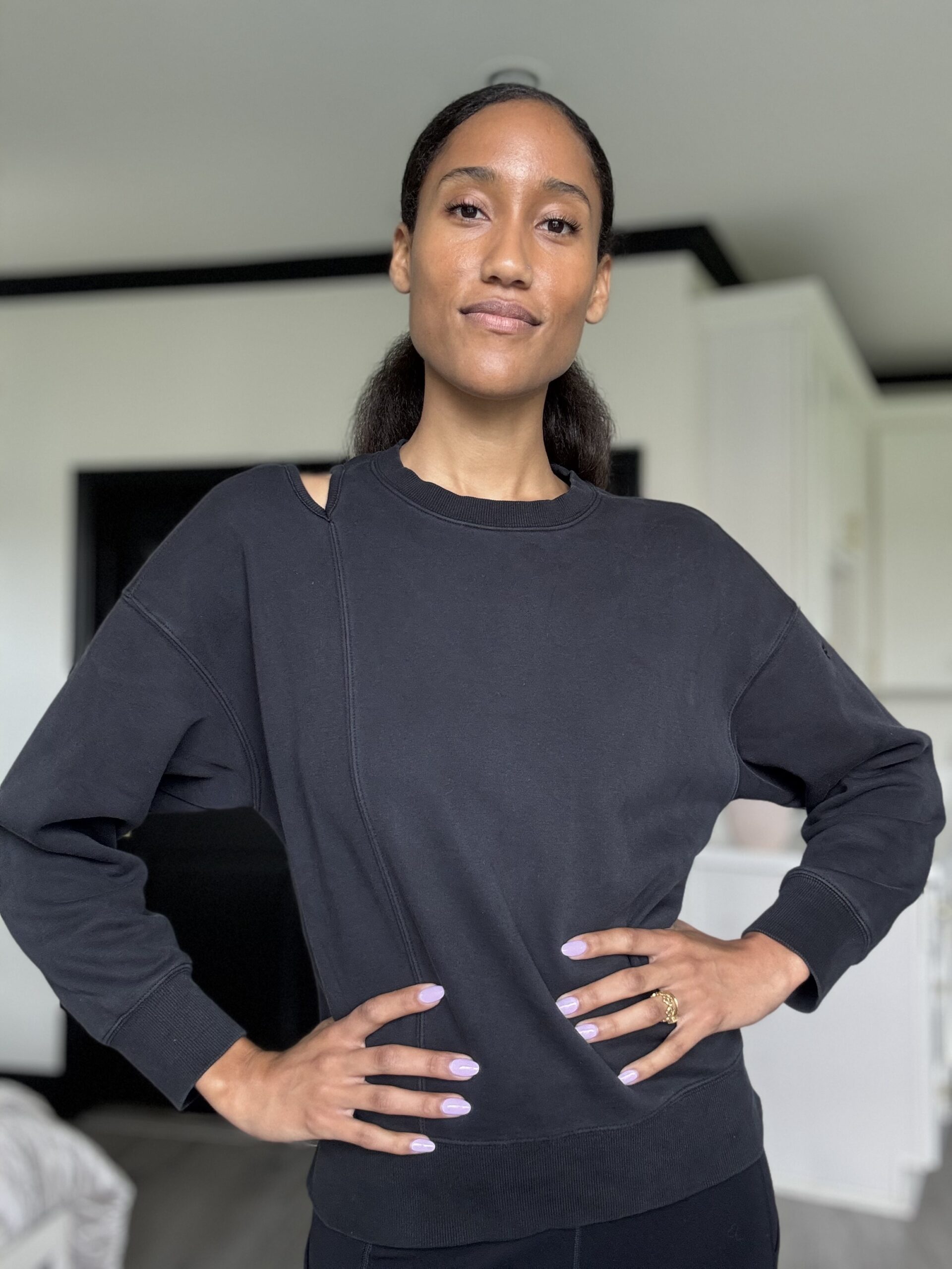 A woman stands indoors with her hands on her hips, wearing a black sweatshirt with a cut-out detail near the collar. She has light purple nails and a neutral expression.