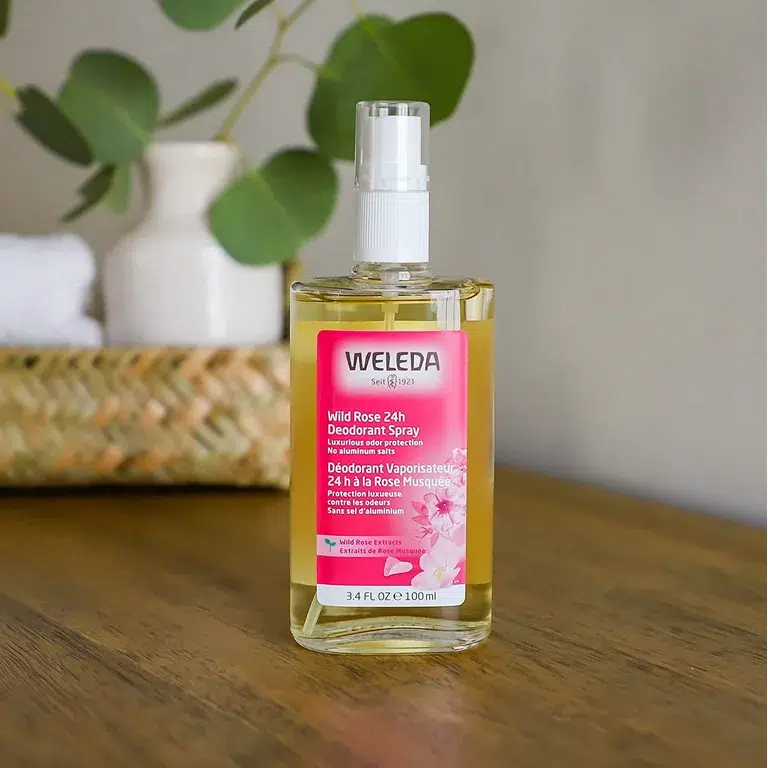 A bottle of Weleda Wild Rose 24h Deodorant Spray sits on a wooden surface with a basket and green plant blurred in the background.