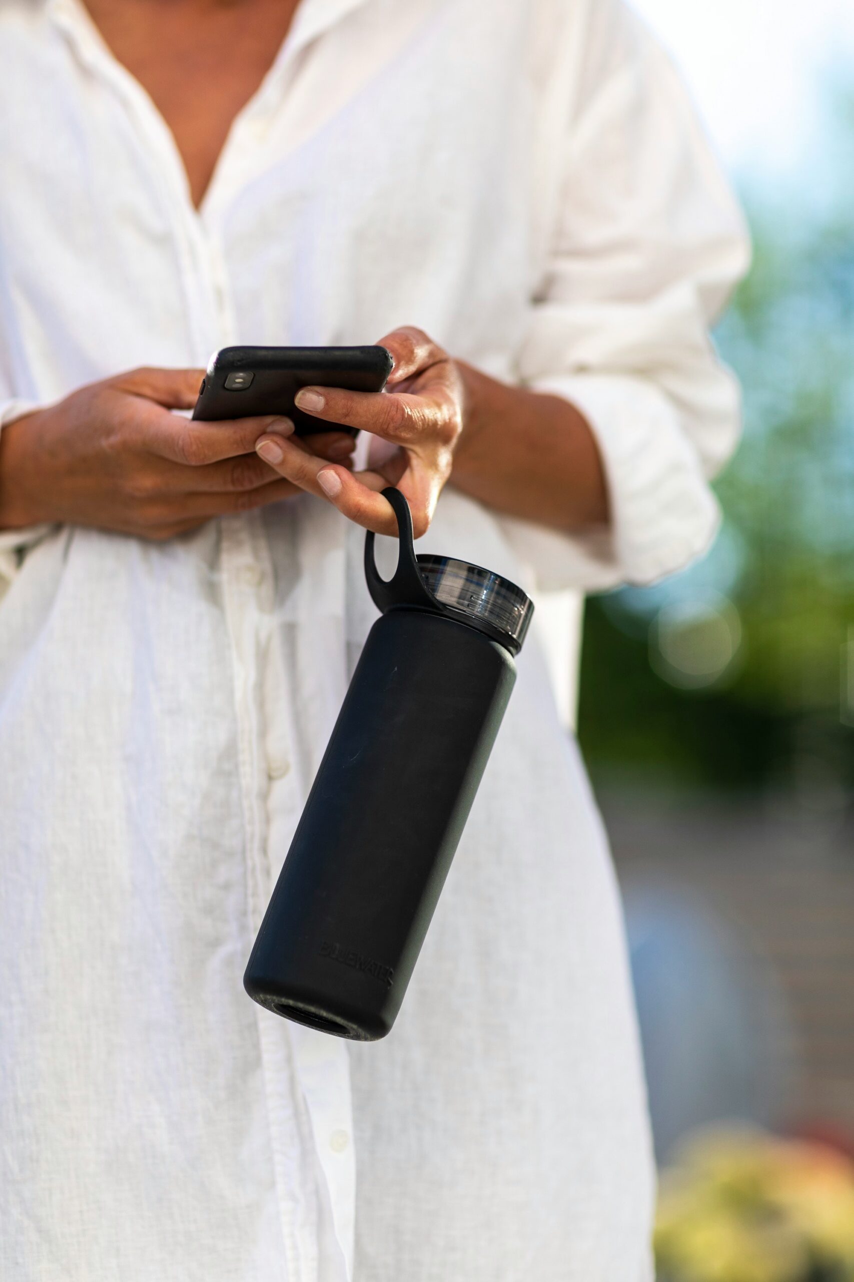 A person wearing a white shirt holds a black smartphone in one hand and a black water bottle in the other, standing outdoors.
