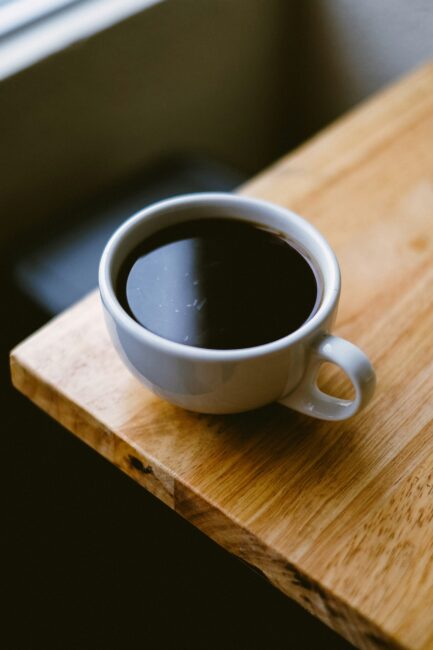A white ceramic cup filled with black coffee sits on a light wooden table near a window.